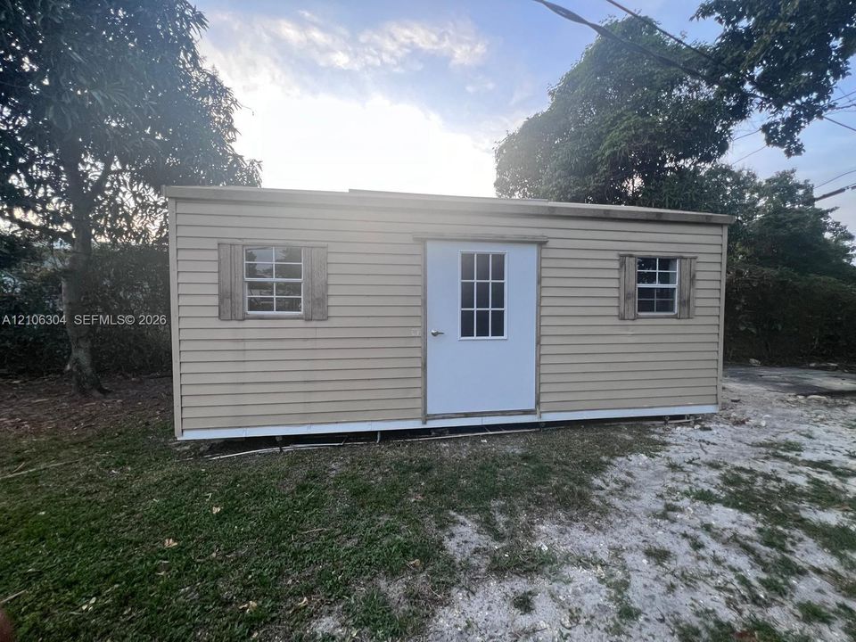 Outbuilding featuring horizontal lap siding, a central entry door with window pane detailing, two exterior windows with wood-tone shutters, and a flat roofline