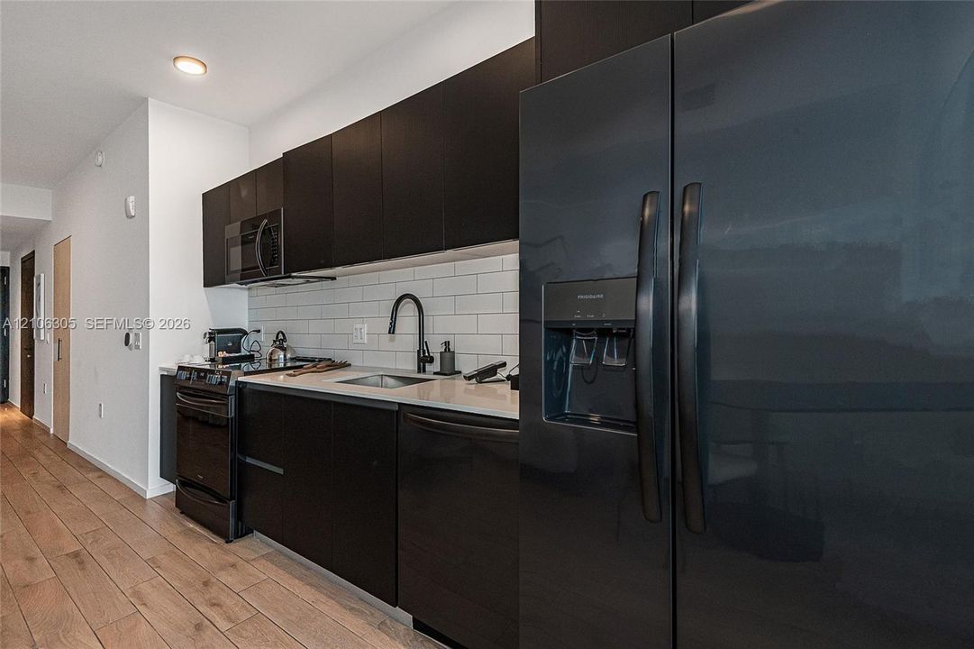 Kitchen featuring dark cabinetry, solid-surface countertops, subway tile backsplash, black appliances, and wood-finish flooring