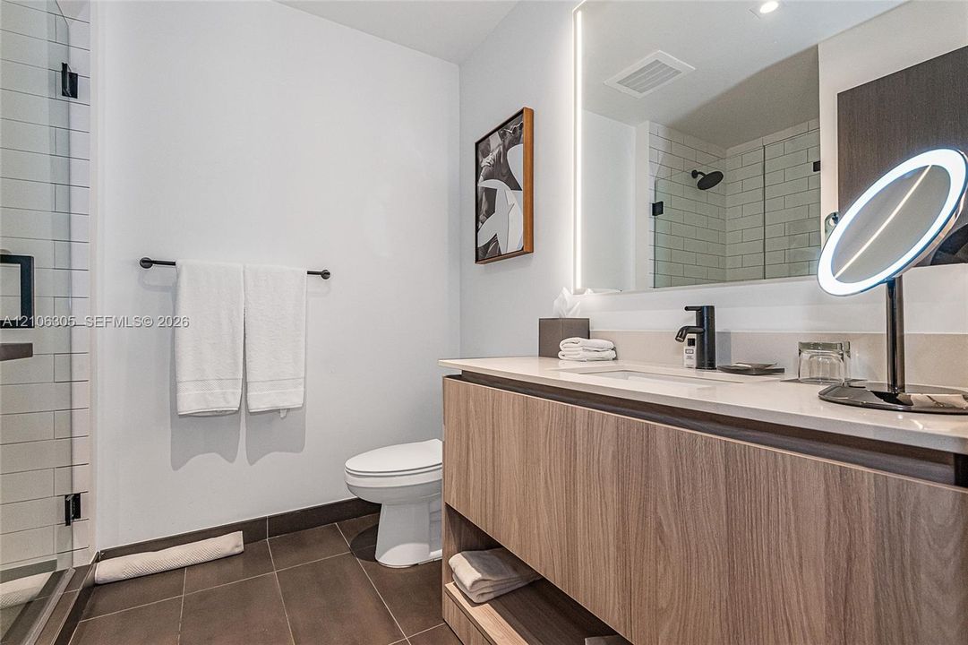 Bathroom featuring a wood-finish floating vanity with a light-tone solid surface countertop, backlit mirror, matte black plumbing fixtures, and tile flooring