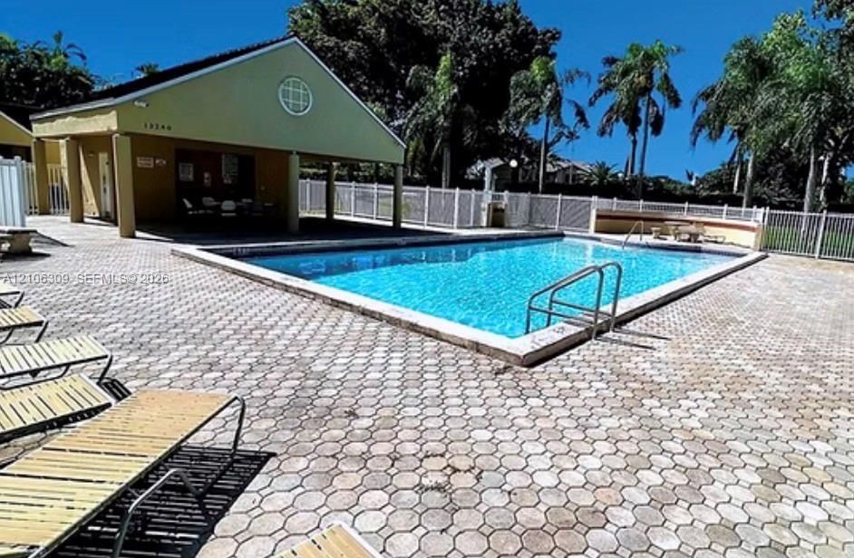In-ground swimming pool with metal ladder entry, surrounded by a paved deck featuring stone-style pavers, enclosed by a perimeter fence, and accompanied by a covered pavilion structure