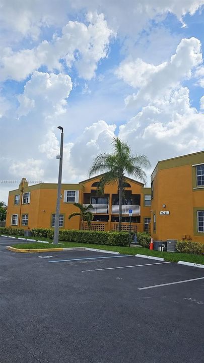 Multi-story residential building featuring stucco exterior, second-level balconies with railing, paved parking area, and manicured perimeter landscaping