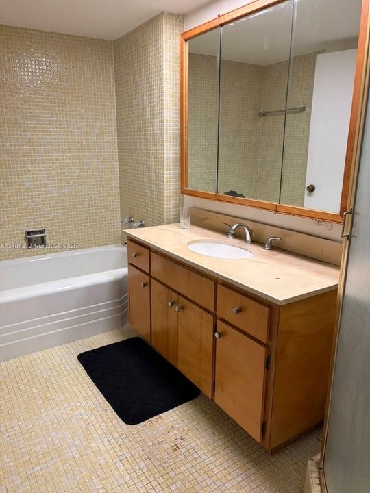 Bathroom featuring a wood-finish vanity with a neutral countertop, undermount oval sink, three-panel medicine cabinet with wood trim, and wall-to-wall mosaic tile detailing around the built-in bathtub