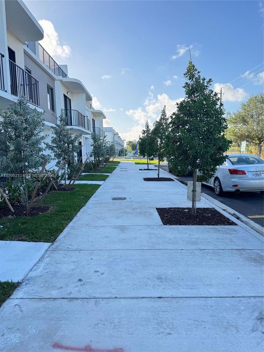 Multi-story exterior featuring a modern facade with rounded architectural accents, integrated metal balcony railings, concrete sidewalk, and landscaped tree-lined beds