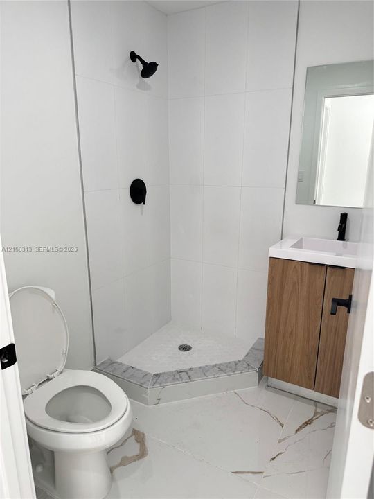 Corner shower with floor-to-ceiling white tile, wood-finish vanity, stone-patterned floor tile, matte black fixtures, and a frameless mirror