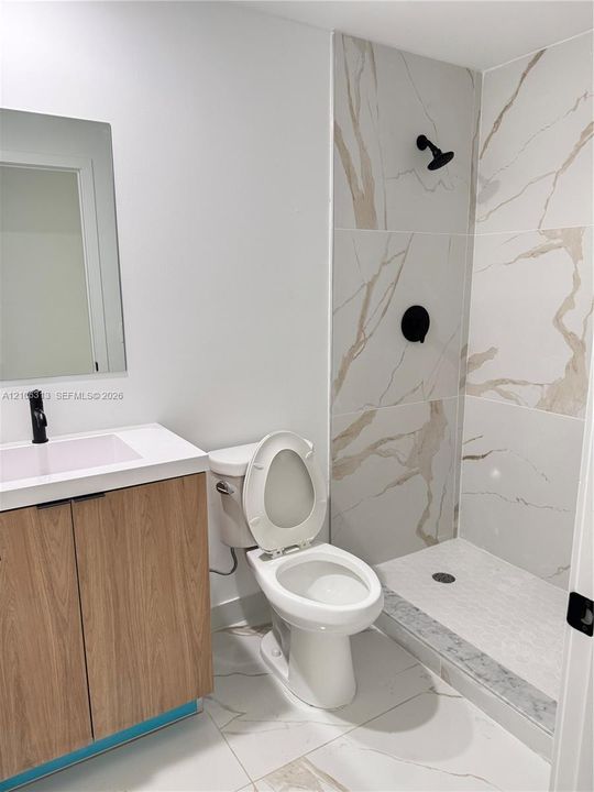 Bathroom featuring a vanity with a light wood-finish cabinet and solid-surface countertop, a frameless mirror, large-format tile flooring, and a walk-in shower with veined wall tile and matte black fixtures