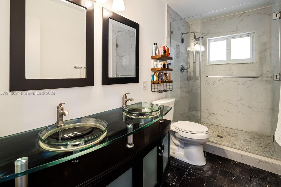 Dual-vanity featuring glass basin sinks, dark-finish cabinetry, frameless glass shower enclosure, and stone-surround wall tiling