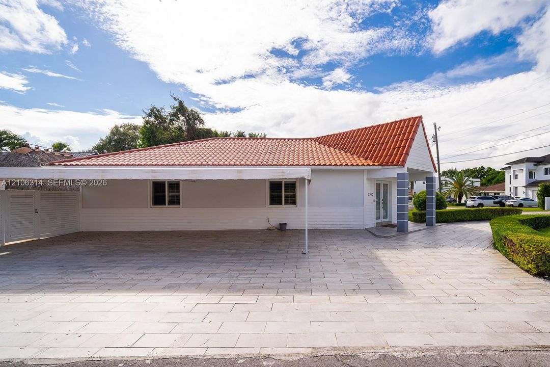 Exterior elevation featuring a terracotta-style tiled roof, a large attached carport with a canopy, light-toned masonry siding, and a paved driveway