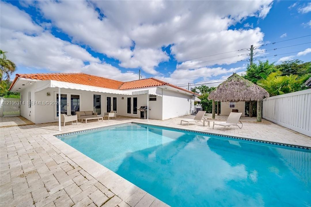 In-ground swimming pool with stone-paver decking, white exterior finish, red-tile roof, and detached thatched-roof gazebo