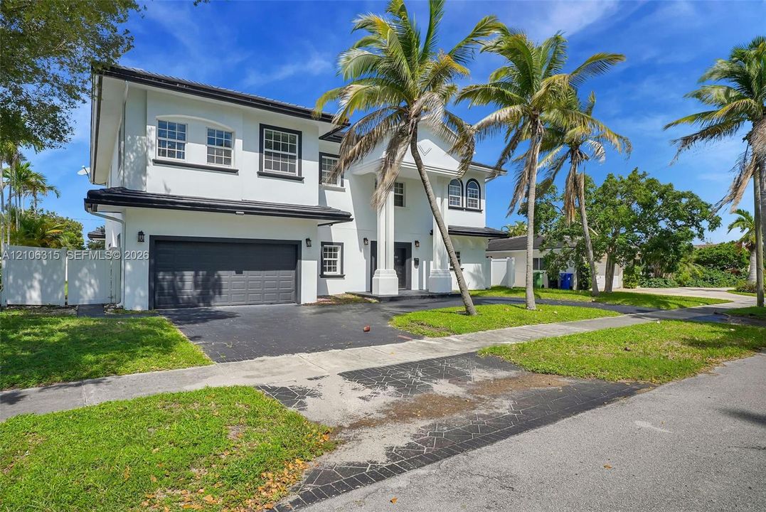 Two-story residence featuring a white exterior, multi-pane windows, a covered entryway with structural columns, an attached two-car garage, and mature palm trees