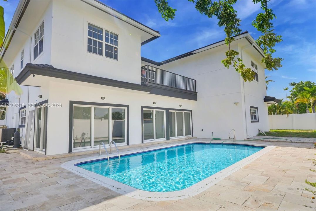Two-story exterior featuring white stucco finish, second-level balcony with metal railing, multiple sliding glass doors, and a rectangular swimming pool surrounded by stone-tile patio
