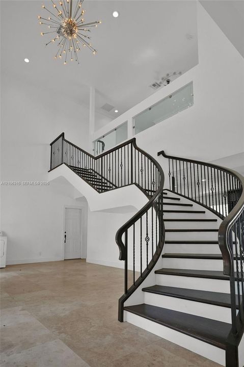 Curved staircase with dark-finished wood treads and metal balusters, polished tile flooring, double-height ceiling, glass-panel balcony railing, and starburst-style chandelier