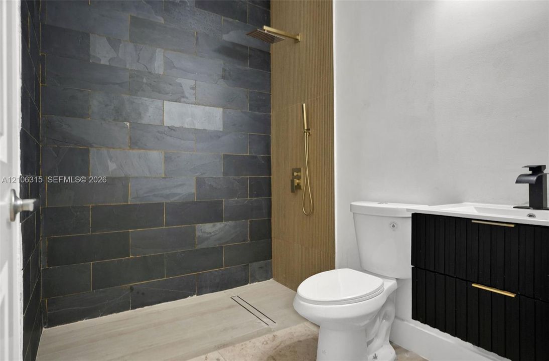 Walk-in shower featuring dark stone-style tile, gold-finish hardware, and a wood-finish panel accent, paired with a black floating vanity and integrated sink