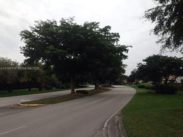 Paved street featuring a landscaped median, concrete curbing, mature trees, and grass lawns