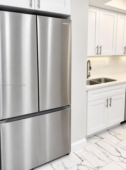 Stainless steel French door refrigerator, white shaker-style cabinetry with brushed metal hardware, solid-surface countertops, dual-basin stainless steel sink with high-arch faucet, and patterned tile flooring