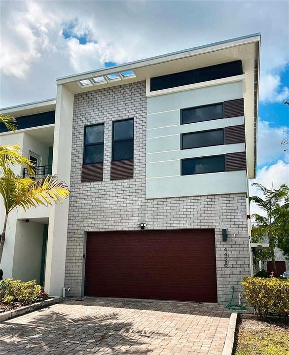 Modern two-story architectural exterior featuring a combination of light-toned brick masonry, horizontal panel siding, a single-car garage with wood-finish door, clerestory windows, and a paved driveway
