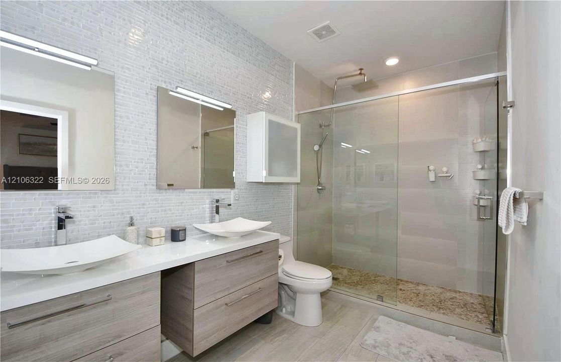 Dual floating vanity with rectangular vessel sinks, chrome faucets, wall-mounted medicine cabinet, and a walk-in glass-enclosed shower featuring a textured stone-finish floor