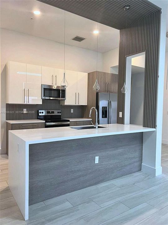 Kitchen featuring a central island with a waterfall-edge quartz countertop, two-tone flat-panel cabinetry, vertical wood-slat accent wall, stainless steel appliances, and wood-finish tile flooring
