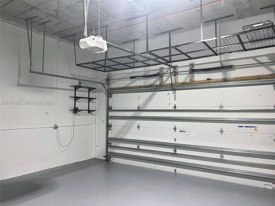 Garage space featuring finished epoxy flooring, white paneled walls, exposed concrete ceiling, integrated overhead metal storage racks, and wall-mounted metal shelving