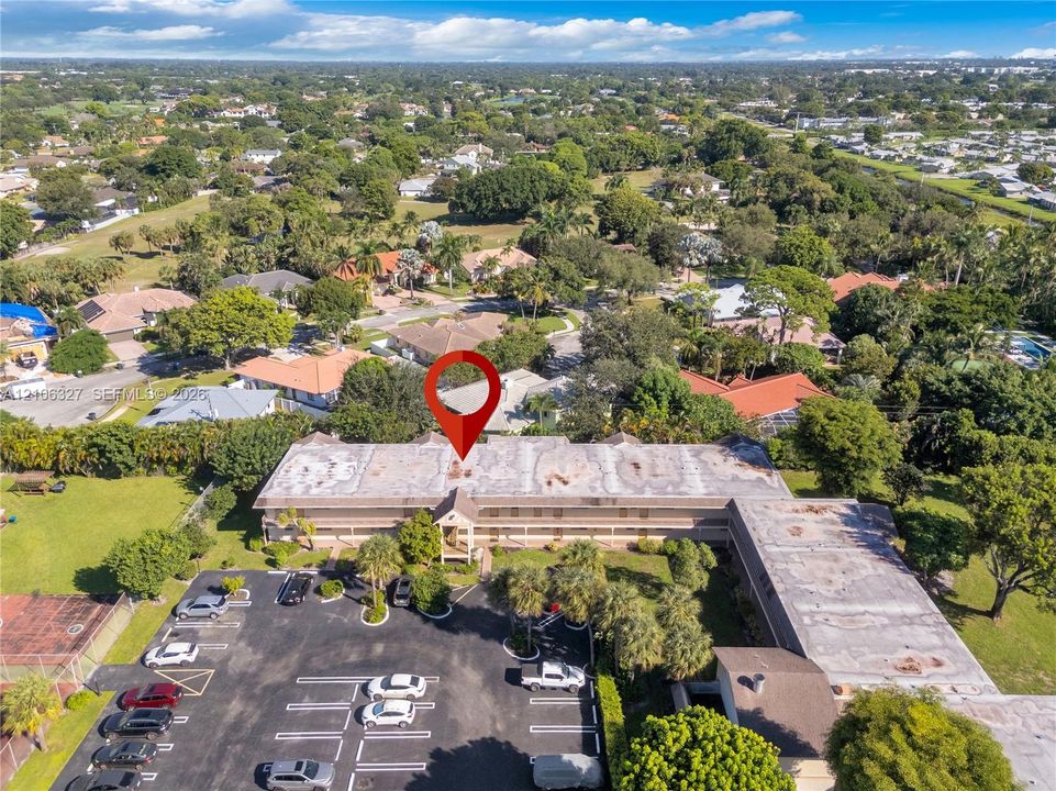 Aerial view of multi-story building featuring an asphalt parking lot, manicured landscaping with mature trees, and ground-level perimeter sidewalk