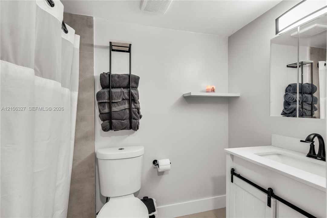 Bathroom featuring a white vanity with a composite countertop, matte black faucet, mirrored medicine cabinet, wall-mounted shelving, and a tiled shower surround