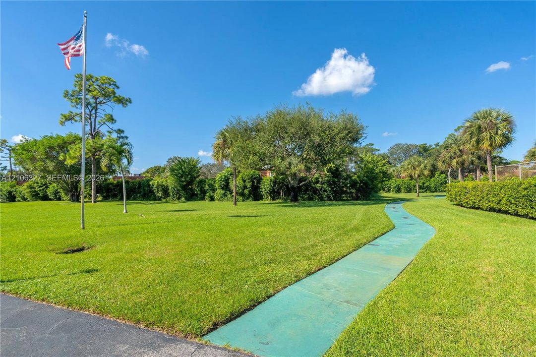 Expansive grounds featuring a manicured lawn, mature trees, ornamental hedges, a paved walking path, and a permanent flagpole