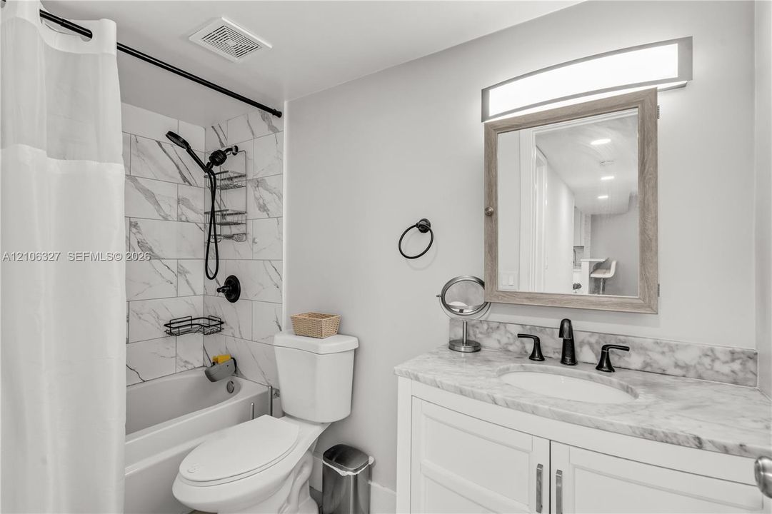 Bathroom featuring a stone-finish countertop with an integrated oval basin, white shaker-style cabinetry, a light-toned wood-frame medicine cabinet, and a bathtub enclosure with marble-patterned wall tile and a matte black shower fixture