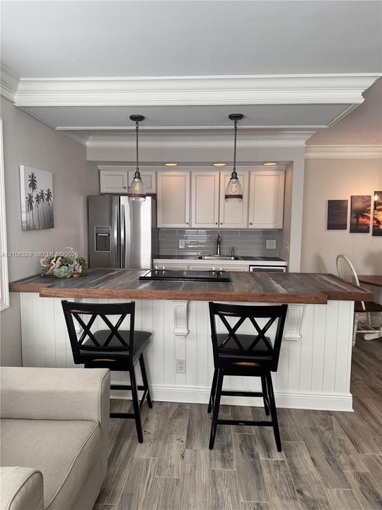 Kitchen featuring a wood-topped peninsula with integrated cooktop, white cabinetry, subway tile backsplash, stainless steel refrigerator, and dual pendant lighting under crown-molded tray ceiling details