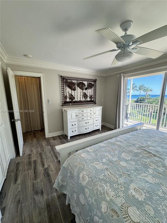 Wood-finish flooring, ceiling fan with integrated light, crown molding, and sliding glass doors opening to a balcony with an ocean backdrop