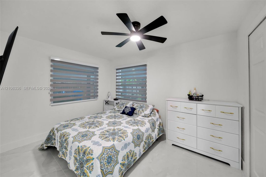 Neutral-toned bedroom featuring large-format tile flooring, a matte black multi-blade ceiling fan with integrated LED lighting, and dual windows equipped with horizontal zebra-style blinds