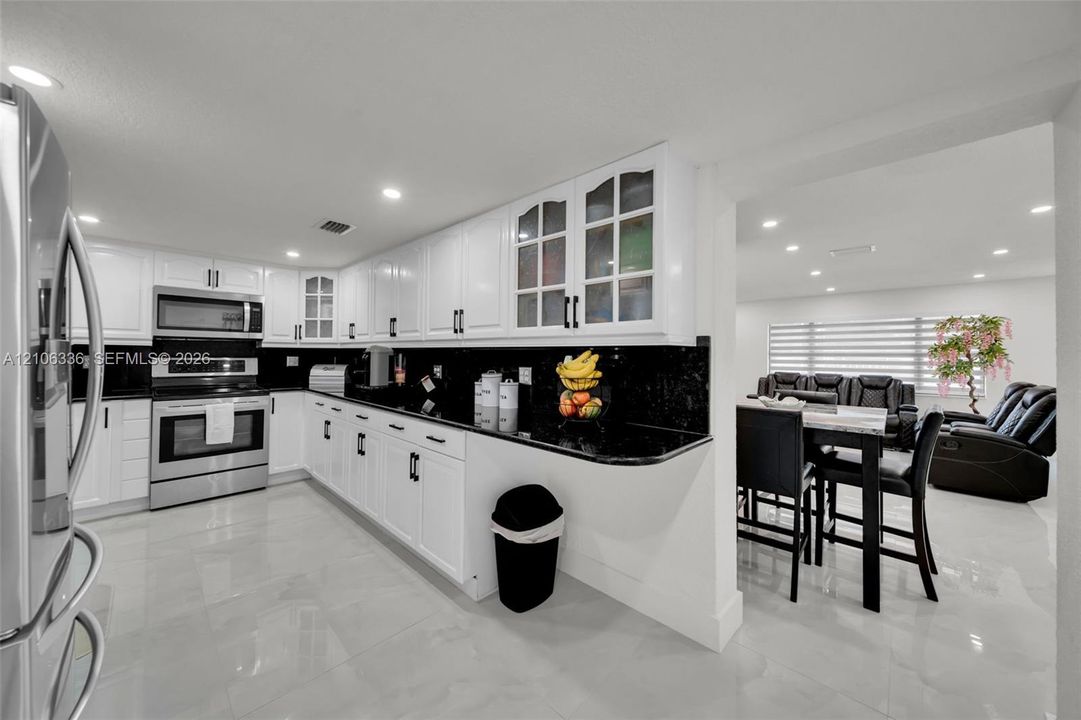 Kitchen featuring white shaker-style cabinetry, black stone countertops, stainless steel appliances, glass-front upper cabinet doors, and polished large-format tile flooring