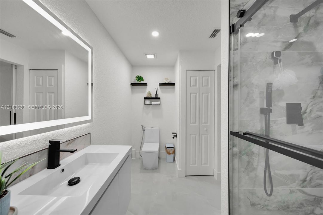 Modern vanity with integrated rectangular sink and matte black faucet, illuminated LED mirror, large-format tile flooring, walk-in shower with glass enclosure, and stone-finish wall tile