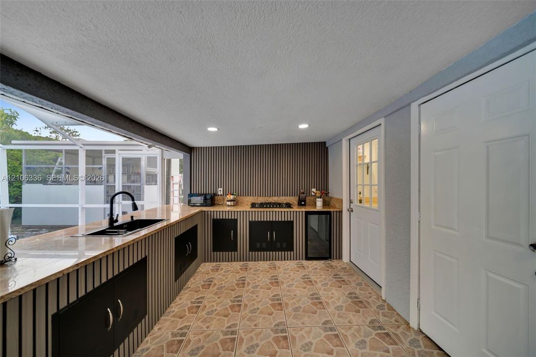 Outdoor kitchen featuring stone-style tile flooring, built-in cabinetry with stone countertops, a drop-in sink with a high-arch faucet, a gas cooktop, and a glass-door beverage cooler