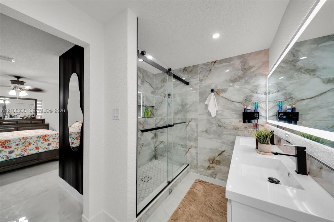 Walk-in shower with floor-to-ceiling large-format tile surround and frameless glass door, dual-sink vanity with matte black faucets, and integrated LED-backlit mirror