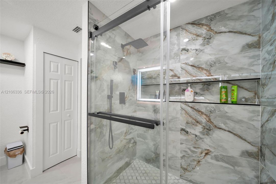 Walk-in shower featuring floor-to-ceiling marbled tile, a built-in recessed niche, rainfall showerhead with handheld sprayer, glass sliding door with matte black hardware, and mosaic-tiled flooring