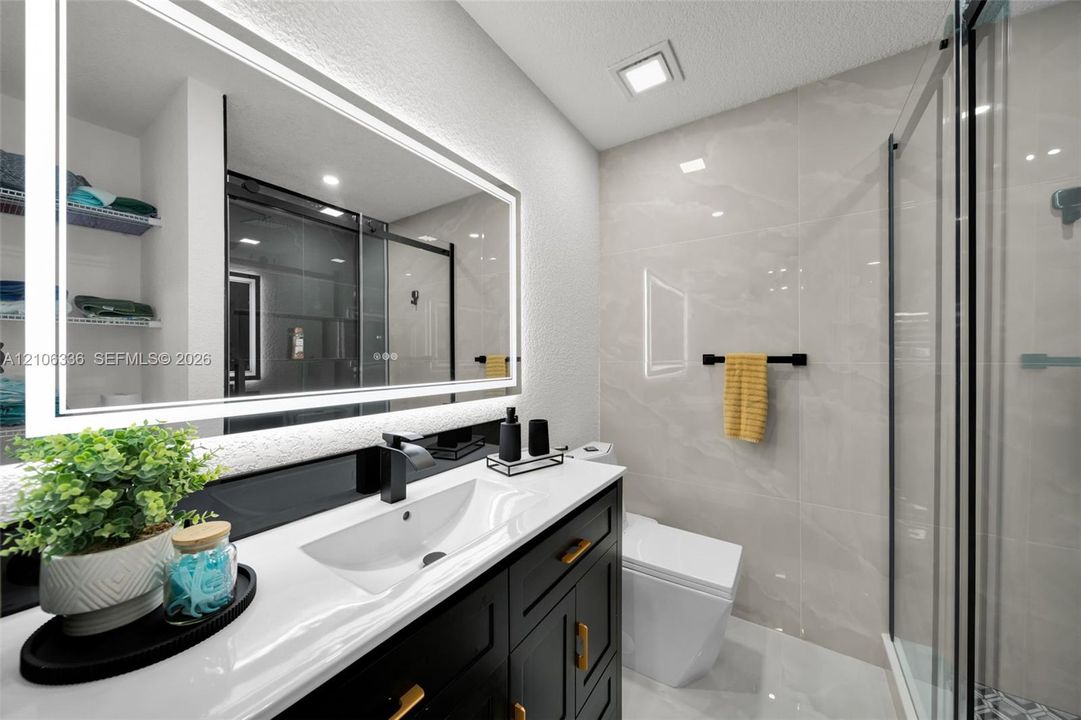 Bathroom vanity featuring a dark-finish cabinet with gold hardware, a white integrated basin, a polished countertop, an illuminated mirror, and a walk-in shower with large-format tile surround