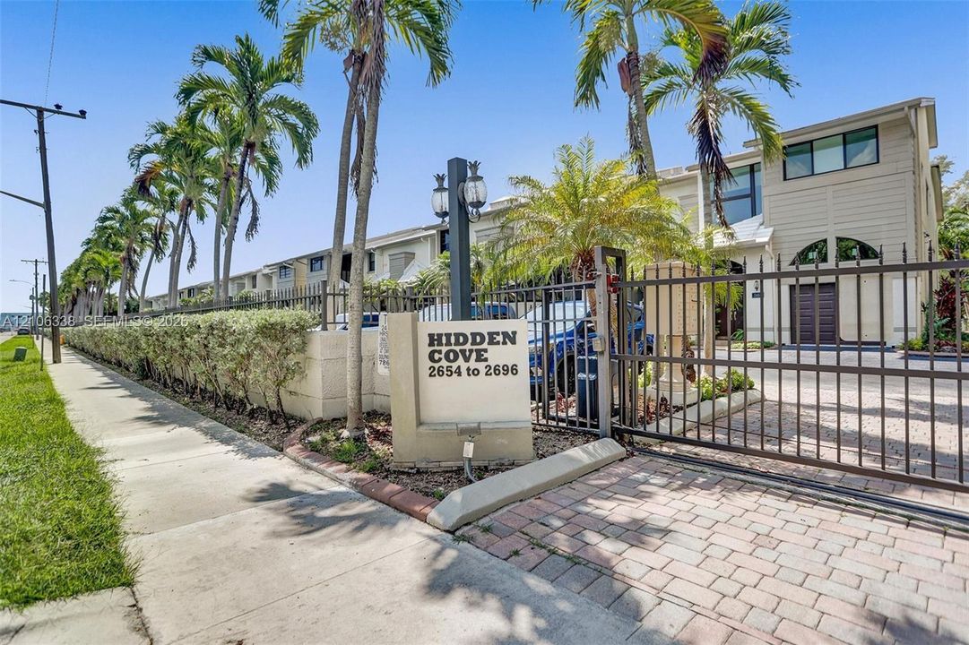 Gated entrance featuring a metal perimeter fence, paver driveway, signage, and mature palm trees