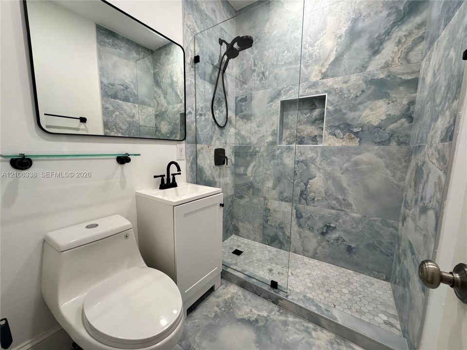 First Floor. Walk-in shower featuring large-format patterned stone-finish tile, glass enclosure, and built-in wall niche, paired with a white vanity and matte black hardware