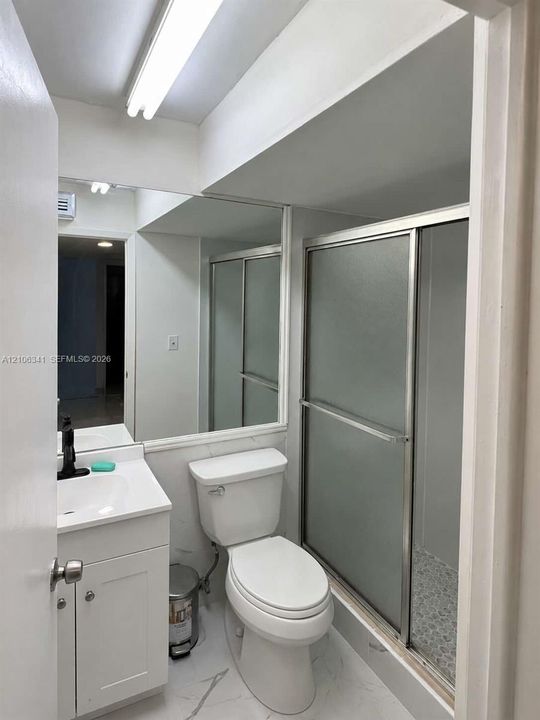 White vanity with a rectangular sink, matte black faucet, wall-spanning mirror, and shower enclosure with frosted glass doors, complemented by stone-pattern tile flooring