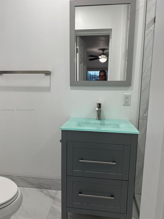 Modern vanity featuring a grey cabinet, a translucent integrated sink, a single-hole chrome faucet, a matching framed mirror, and light-toned tile flooring