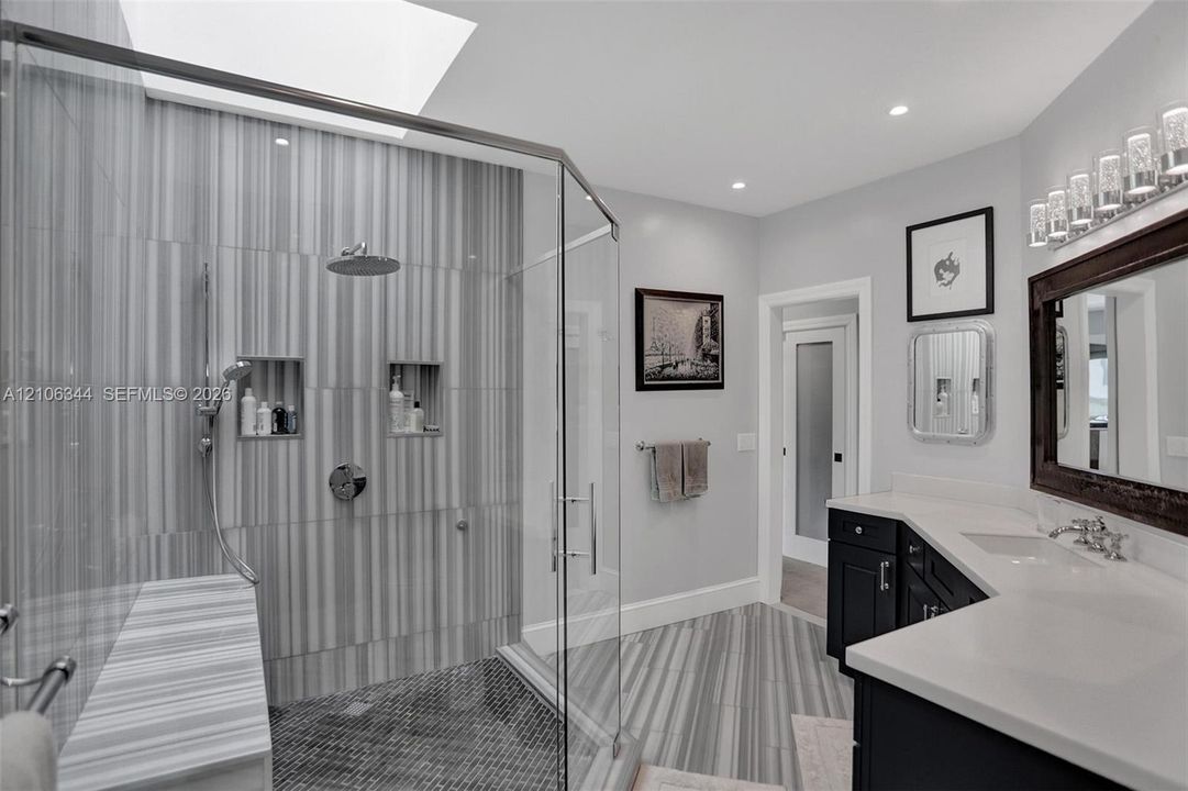 Walk-in shower with vertical tile surround, dual recessed shower niches, overhead rainfall showerhead, glass enclosure, and stone-finish bench seat, accompanied by a dual-sink vanity with solid-surface countertops