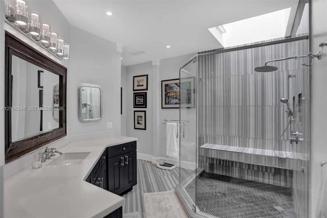 Bathroom featuring a glass-enclosed shower with a built-in bench, dual-sink vanity with a solid-surface countertop, dark cabinetry, and a skylight