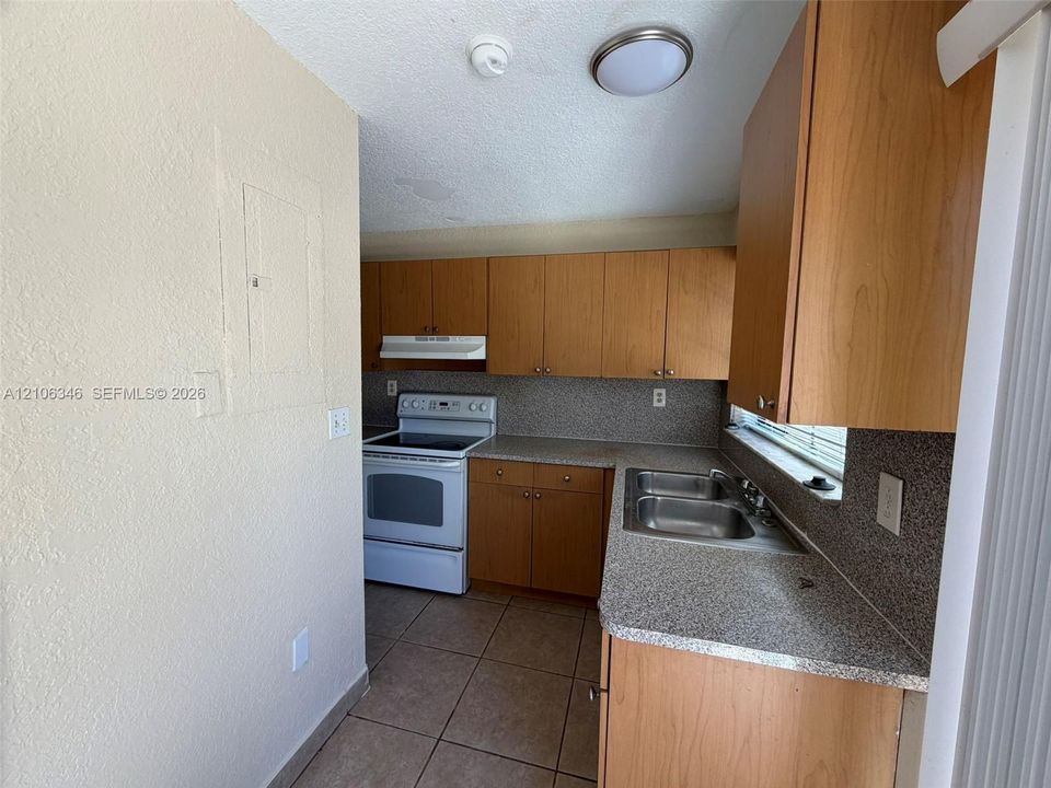 Kitchen featuring wood-finish cabinetry, stone-texture laminate countertops, stainless steel double-basin sink, electric range with overhead vent hood, and tile flooring