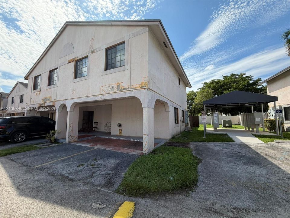 Two-story building exterior featuring a covered ground-level entryway, stucco facade, and an attached carport