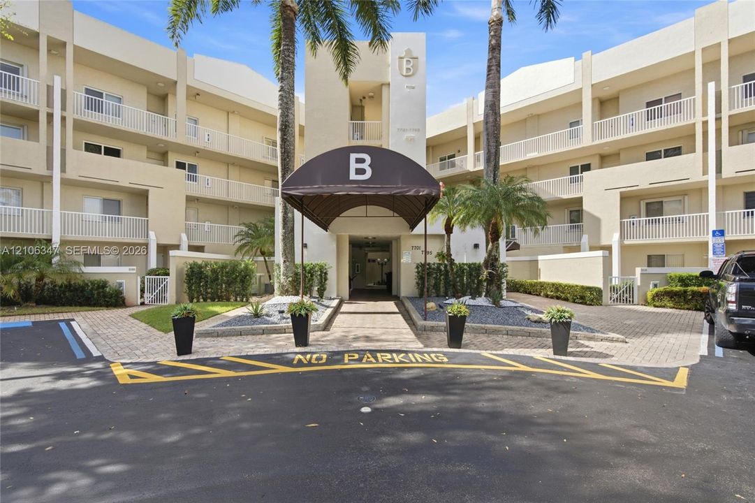 Multi-story building exterior featuring a covered entryway with awning, integrated balconies, paver stone hardscaping, and mature palm landscaping