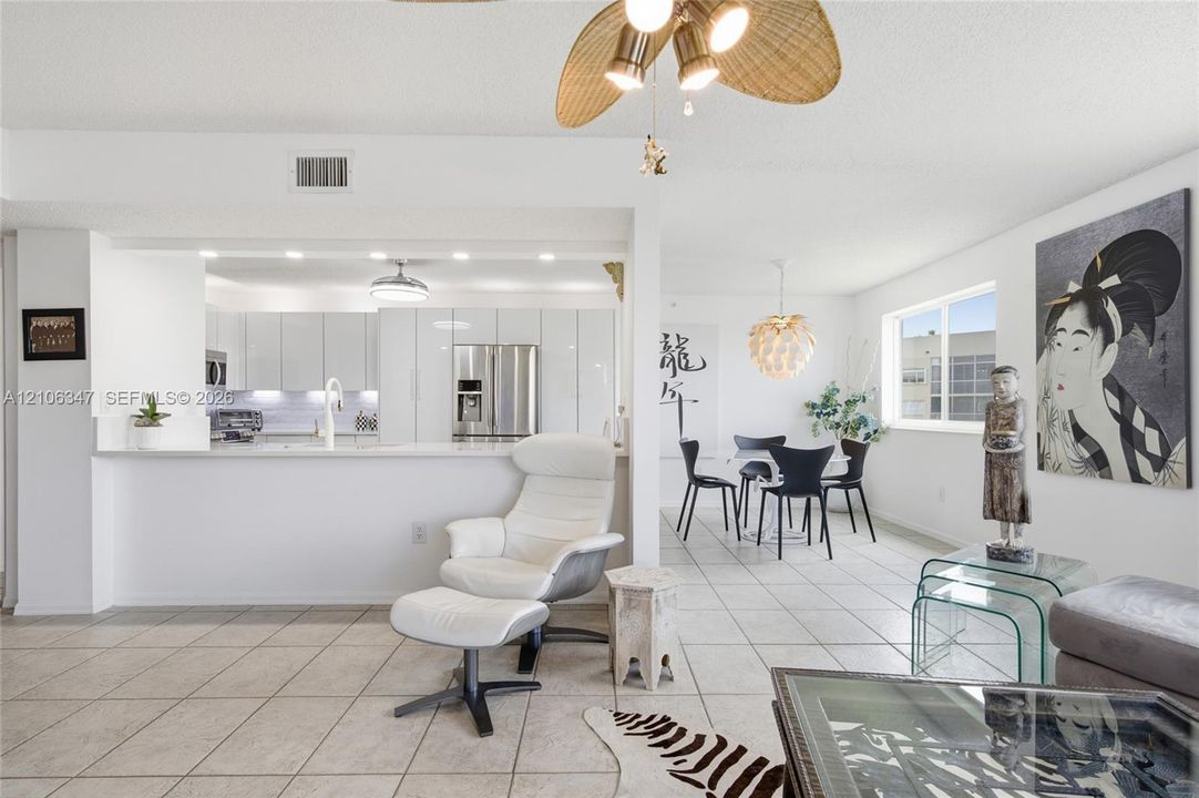 Open-concept interior featuring light-toned tile flooring, white slab-front cabinetry, stainless steel kitchen appliances, a stone-surface breakfast bar, and decorative ceiling fan with woven-finish blades