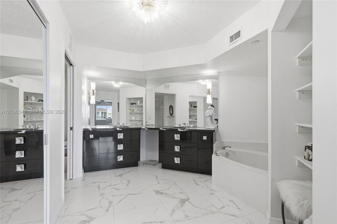 Large-format white tile flooring with grey veining, dual-basin vanity with dark cabinetry, expansive wall-to-wall mirrors, built-in shelving, and a deep soaking tub