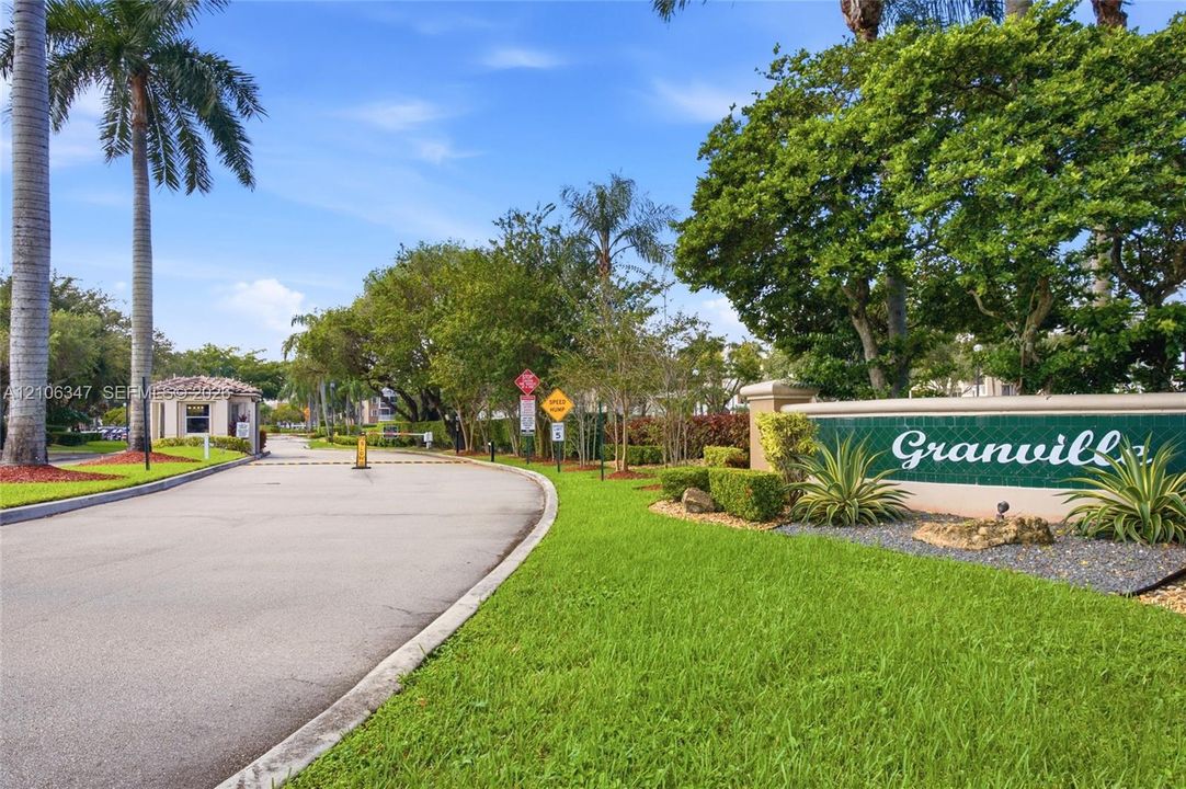 Gated community entrance featuring paved access, a security booth, manicured lawns, mature palm trees, and professional landscaping