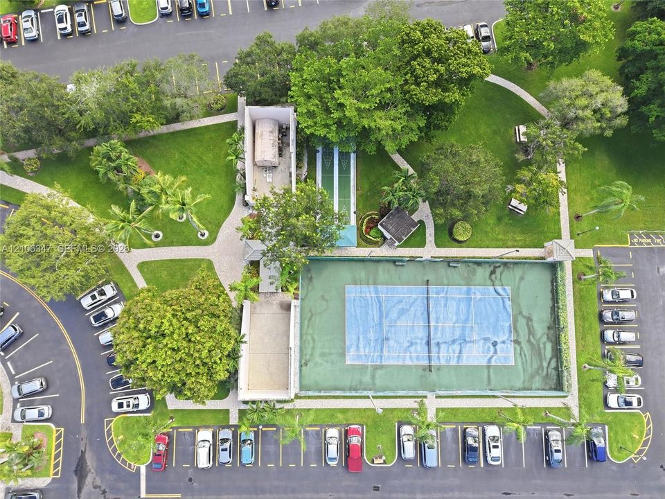 Aerial perspective of recreational grounds featuring a fenced tennis court, shuffleboard court, paved walkways, and extensive landscaped green space
