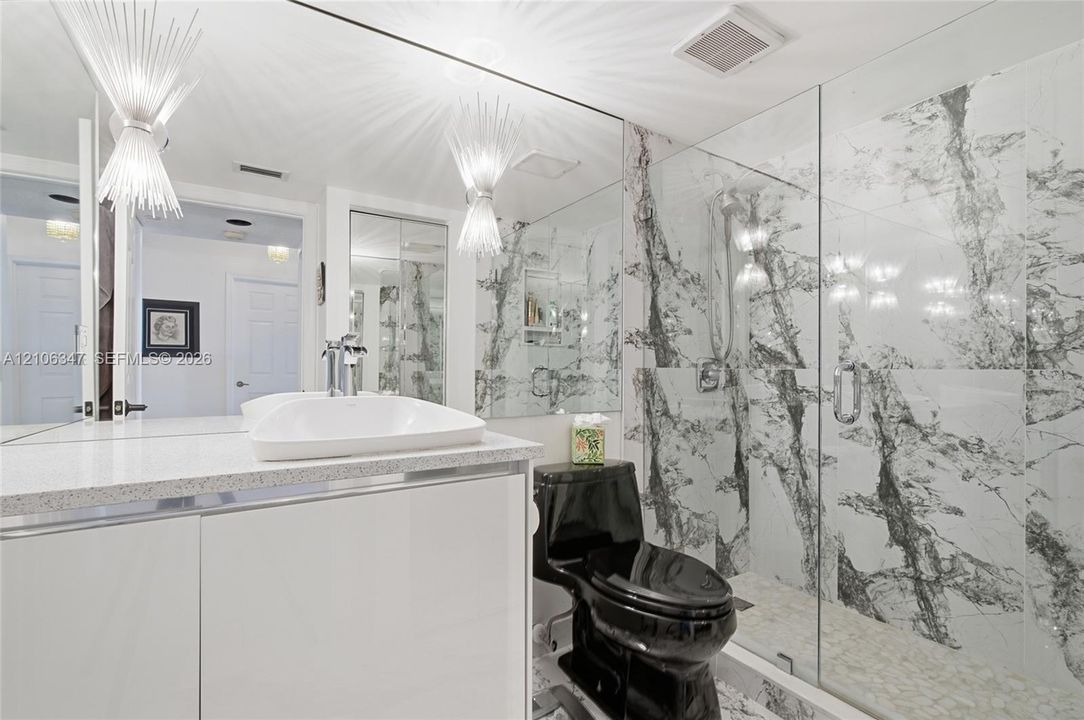 Bathroom featuring floor-to-ceiling veined stone-style tile shower, frameless glass enclosure, glossy white vanity with integrated basin, expansive wall mirror, and dual starburst-style wall-mounted lighting