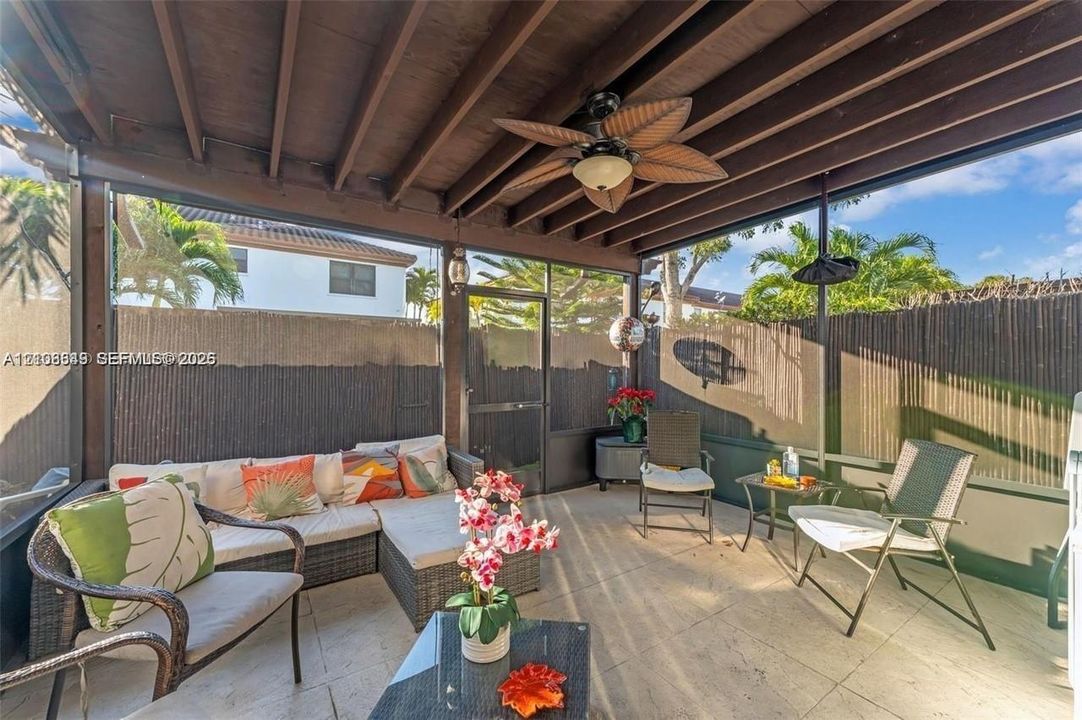 Covered patio with wood-beam ceiling and ceiling fan, concrete flooring, and wrought-iron seating areas overlooking a fenced backyard with mature landscaping and palm trees